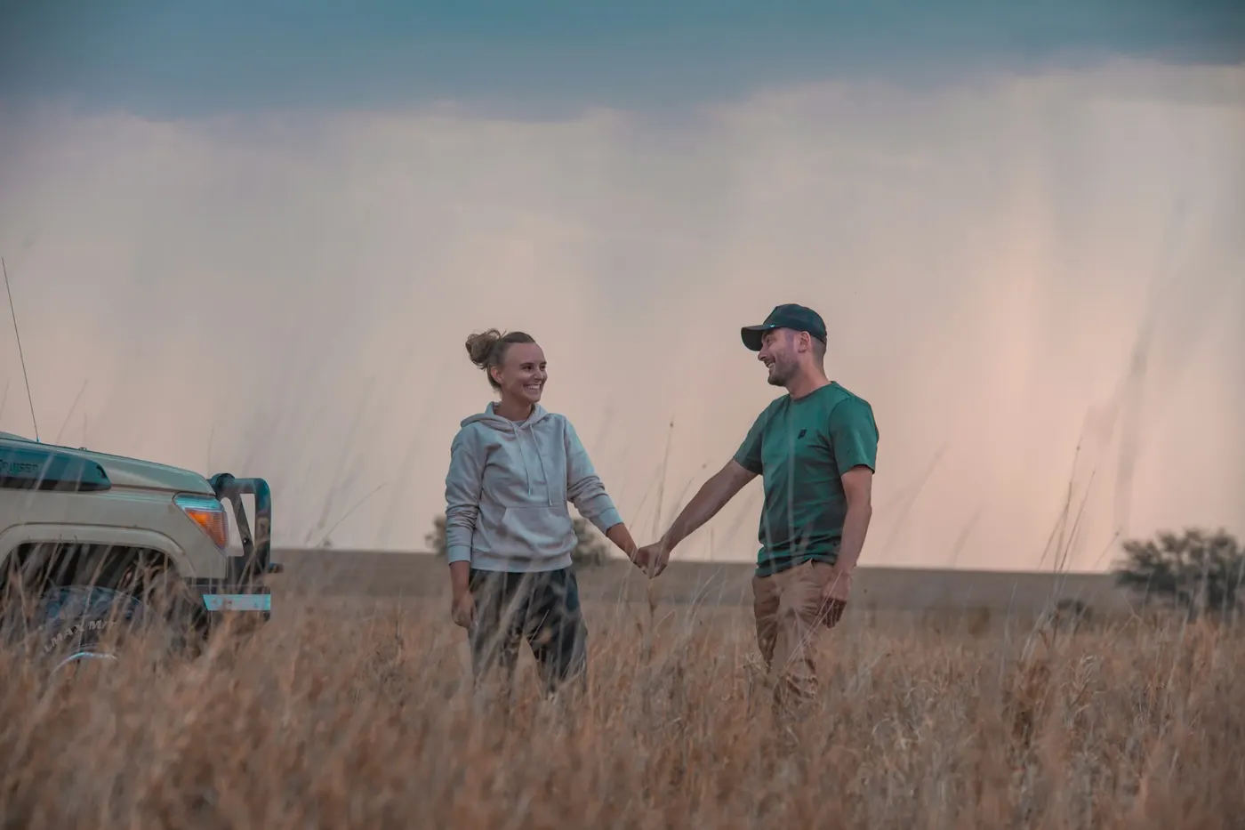 A couple laughing and holding hands in long grass beside a safari vehicle